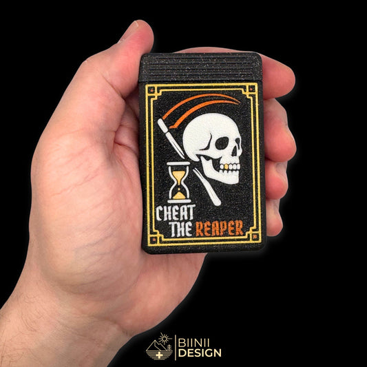 Cheat the Reaper pillbox shown in hand for size reference — compact gothic organizer for vitamins, supplements, or medication.