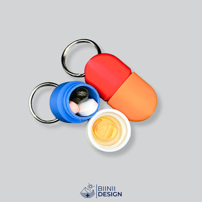 Blau-weiße Compact-Pillendose für den Schlüsselanhänger mit abgeschraubtem Unterteil und Tabletten darin, an eine Mini-Pillendose in Rot und Orange gelehnt.