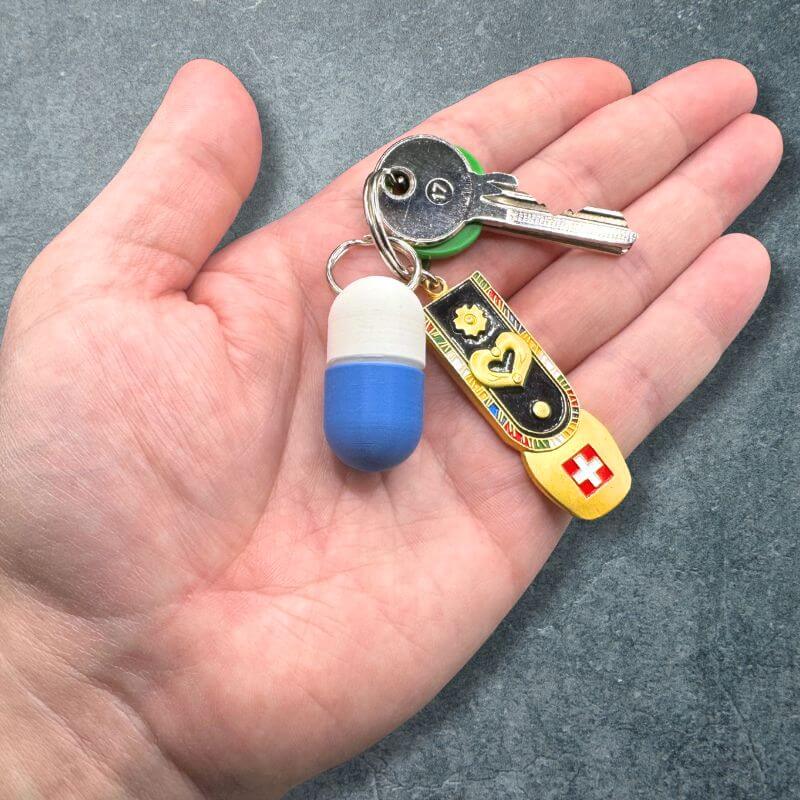 White and blue compact keychain pill holder on a key ring with a key and a Swiss charm, resting in an open palm.