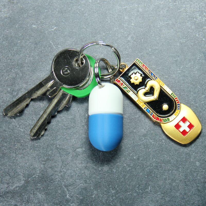 White and blue compact keychain pill holder on a key ring with two keys and a Swiss enamel charm, on slate.