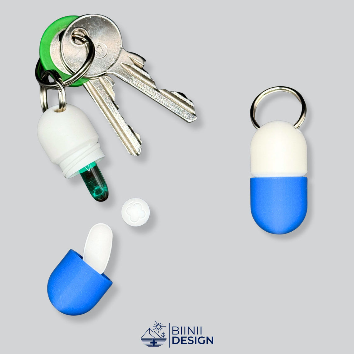 Offene weiß-blaue Schlüsselanhänger-Pillendose in Kapselform am Schlüsselring, mit einer Softgel-Kapsel und einer länglichen Tablette.