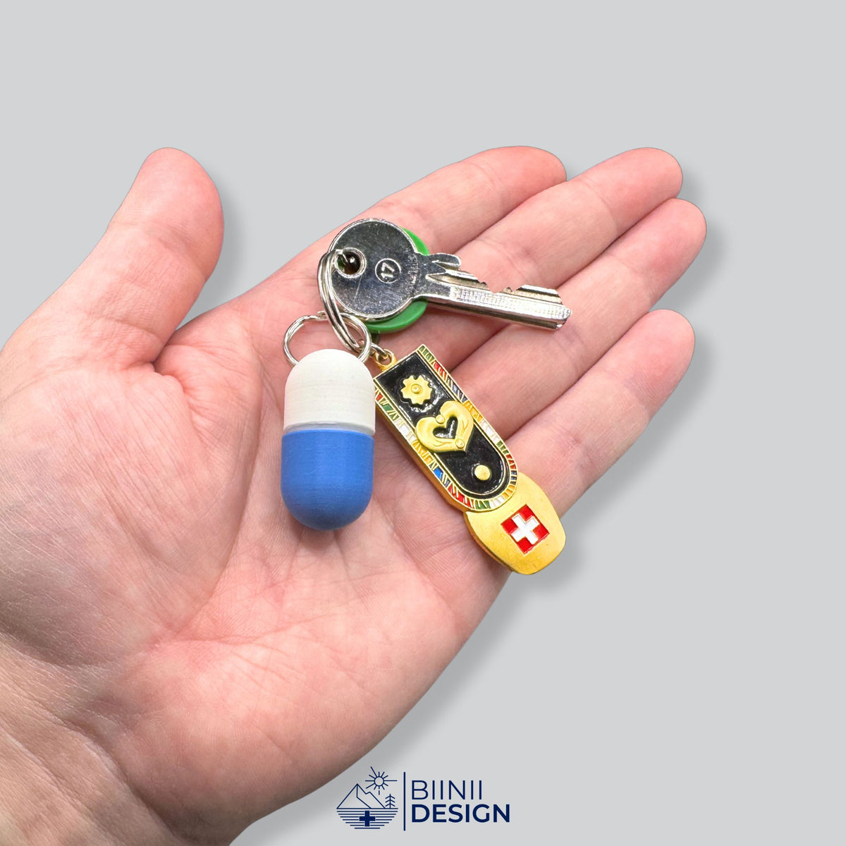 Weiß-blaue kompakte Schlüsselanhänger-Pillendose in einer offenen Hand, an einem Schlüsselring mit Schlüssel und Schweizer Fahnen-Anhänger.