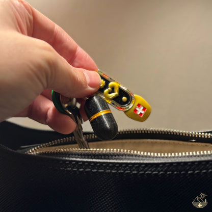 Pilulier porte-clés Deluxe galaxy black à accent doré, un porte-clés avec le drapeau suisse sur l'anneau, glissé à la main dans un sac à main en cuir bleu.