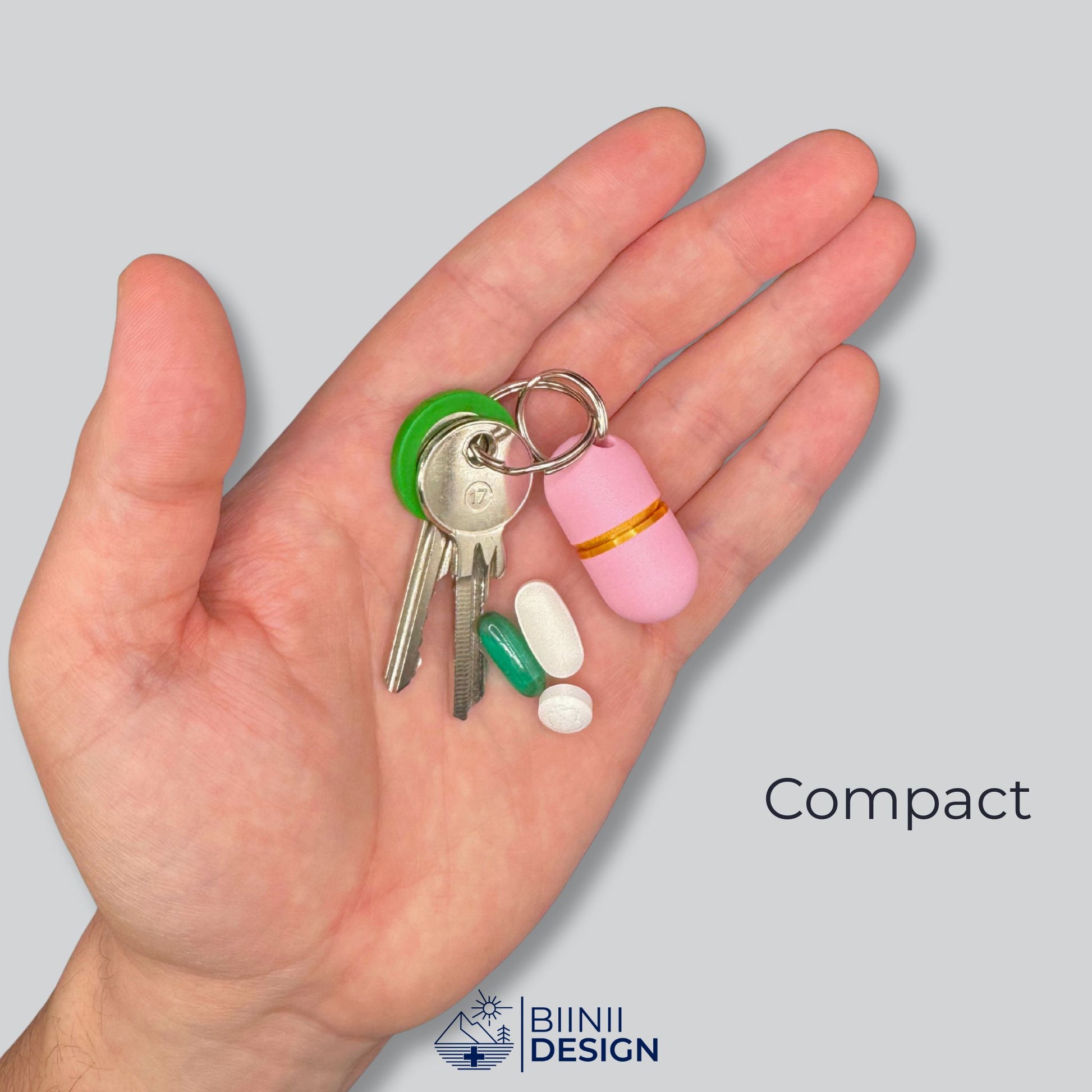 Deluxe compact keychain pill holder (solid pink, gold ring) on keys, held in hand. Discreet pocket pill case for 2–3 daily doses.