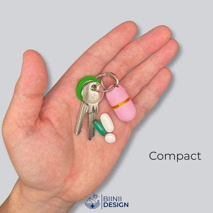 Compact-Deluxe-Pillendose in zartem Rosa mit Gold-Akzent an Schlüsseln in einer offenen Hand, daneben ein Softgel und zwei weiße Tabletten.