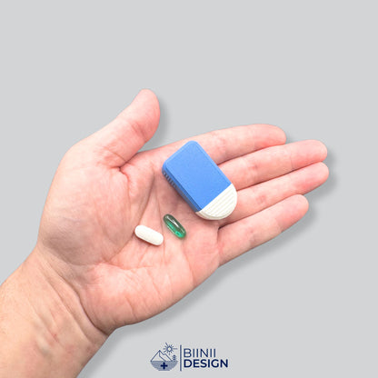 Pilulier compact 2 compartiments bleu et blanc tenu en main pour donner l'échelle, une gélule et un comprimé oblong blanc à côté.