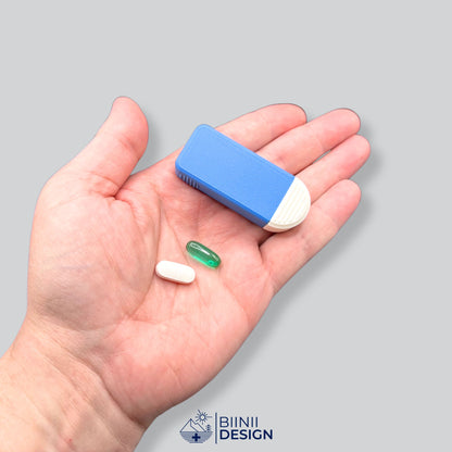 Blue and white compact 3-compartment pill box held in hand for scale, a softgel and a white oblong tablet beside it.