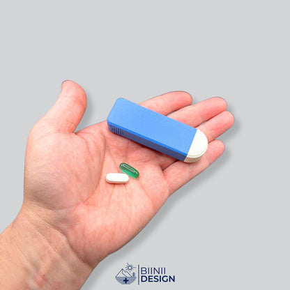 Pilulier compact 4 compartiments bleu et blanc tenu en main pour donner l'échelle, une gélule et un comprimé oblong blanc à côté.