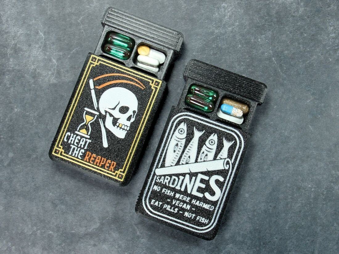 Two black Artsy pill organizers with UV-printed lids, Cheat the Reaper skull and Sardines tin, lids slid open with capsules inside, on slate.