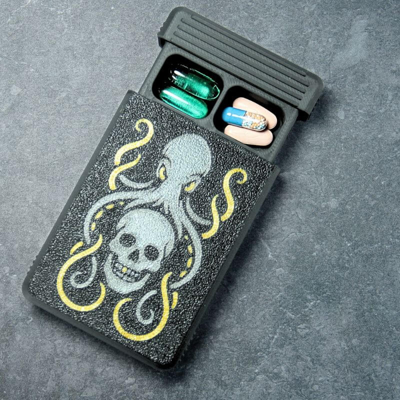 Black Artsy pill organizer, Kraken edition: UV-printed anthracite kraken and skull with gold tentacle accents, lid slid open with capsules inside, on slate.