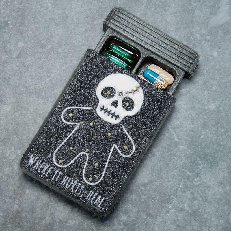 Black Artsy pill organizer, Healing Voodoo edition: UV-printed skull-headed doll with gold-filled pin marks and copper stitches, the words Where it hurts, heal, lid slid open with capsules inside.