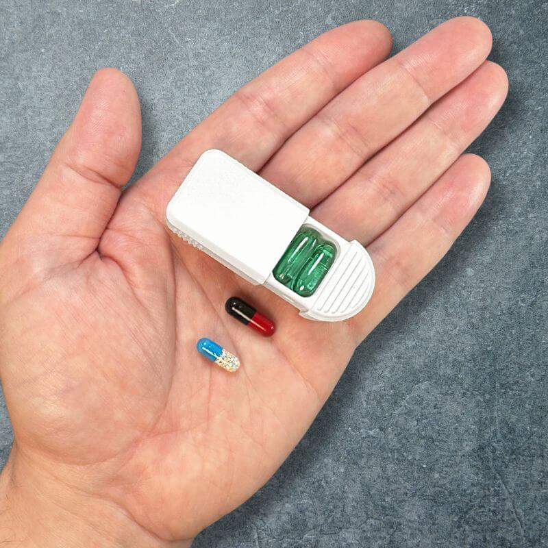White daily pill organizer with the lid slid open, holding two green capsules, in an open palm with two more capsules beside it.