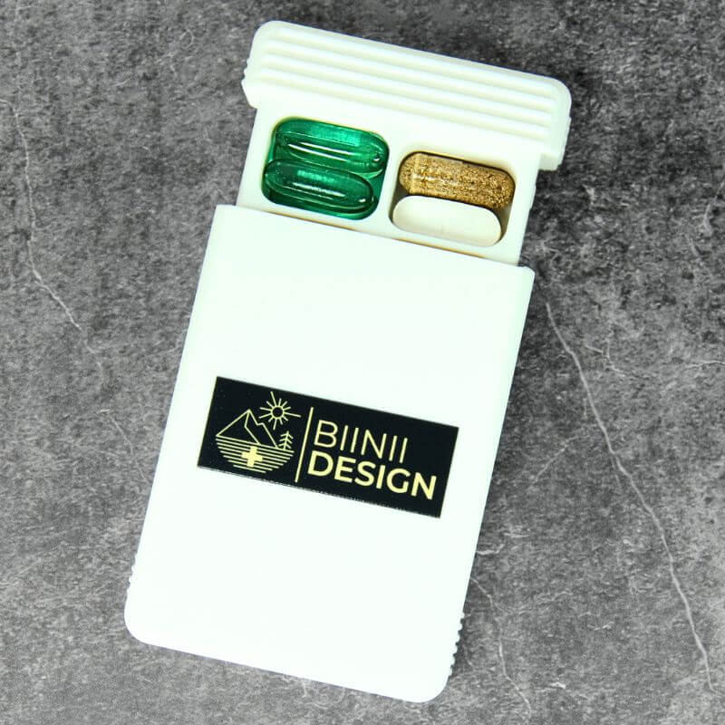 White pill organizer with the Biinii Design logo UV-printed on the lid, an example of a company logo edition, lid slid open with capsules inside, on slate.