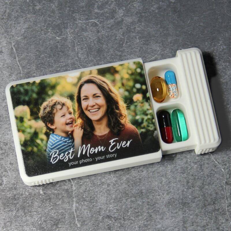 White photo pill organizer, lid UV-printed with a photo of a smiling mother and child and the words Best Mom Ever, your photo, your story, slid open to show capsules in two compartments.