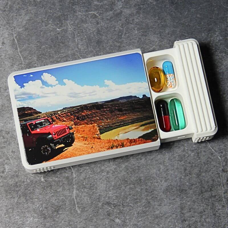 White photo pill organizer, lid UV-printed with a photo of a red off-road car above a desert canyon, slid open to show capsules in two compartments, on slate.