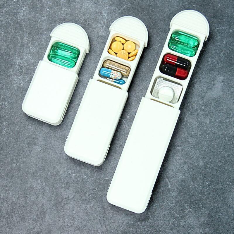 Three white daily pill organizers with the lid slid open, from one to three compartments, filled with capsules and tablets, on slate.