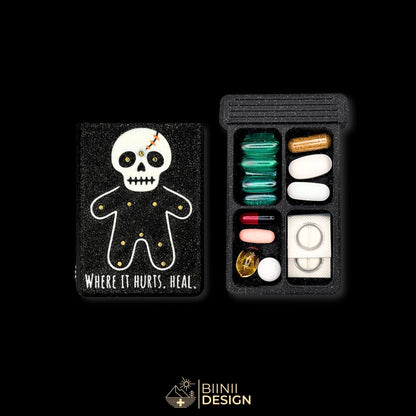 Healing Voodoo art edition pill organizer lid beside its 4-compartment base filled with assorted supplements, stitched voodoo doll artwork.