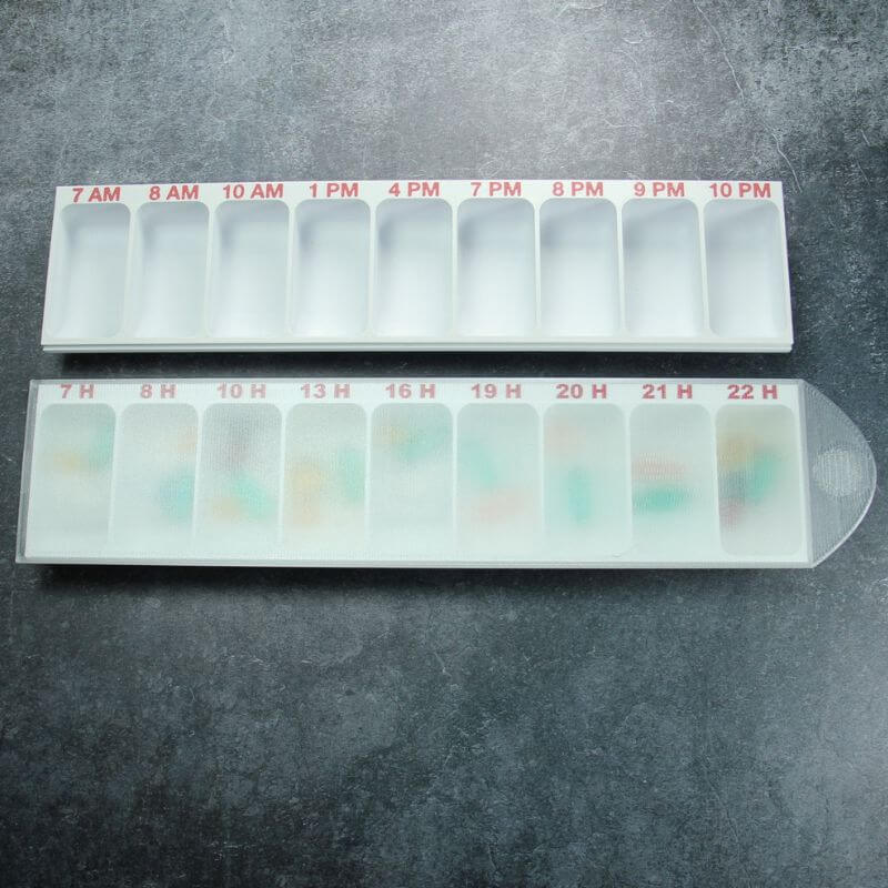 Two white Hourly pill organizers showing the two time formats: 7 AM to 10 PM on the open one, 7 H to 22 H on the closed one with its translucent lid, on slate.