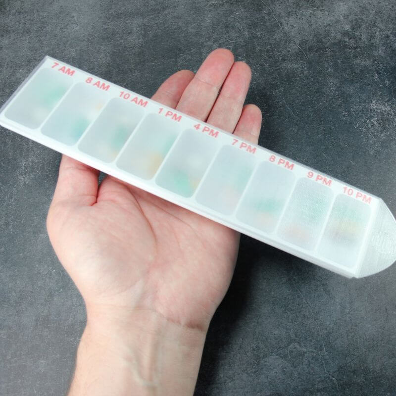 White Hourly pill organizer with nine compartments printed 7 AM to 10 PM, translucent lid closed, held flat in an open hand.