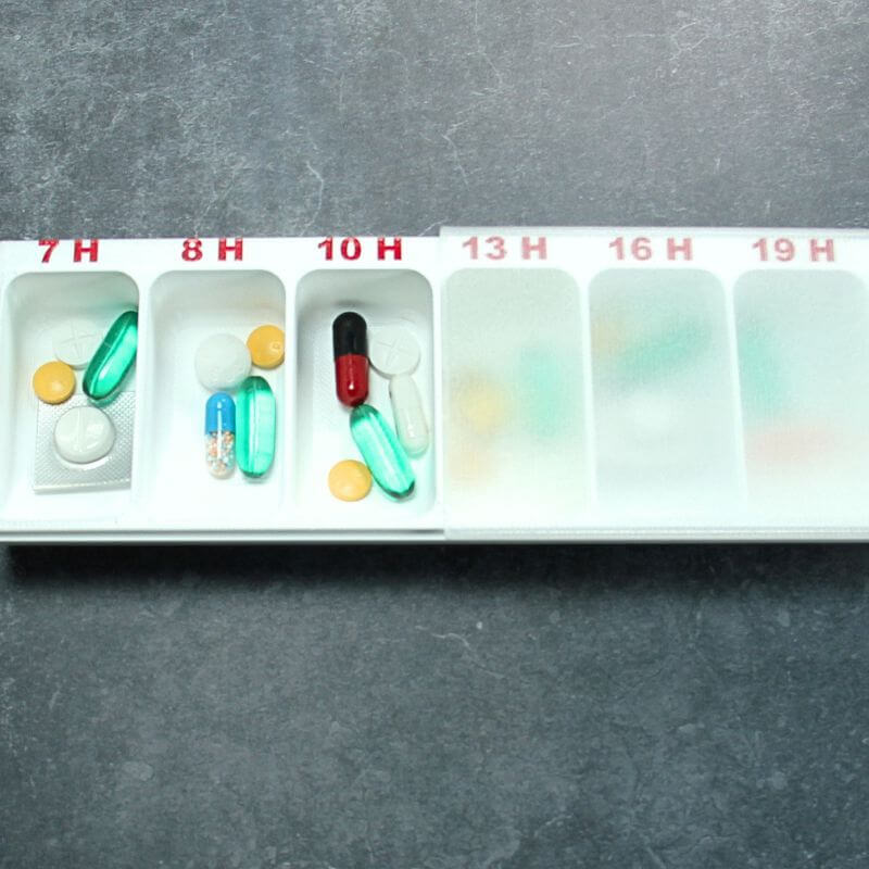 White Hourly pill organizer with compartments printed 7 H to 19 H, translucent lid slid halfway open, morning compartments filled with capsules and tablets, on slate.