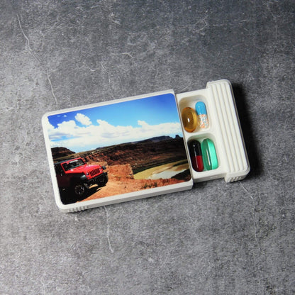 Custom pill organizer with a UV-printed photo of a red 4x4 in a desert canyon on the white lid, lid partly closed to reveal the first row of capsules in the 5-compartment base