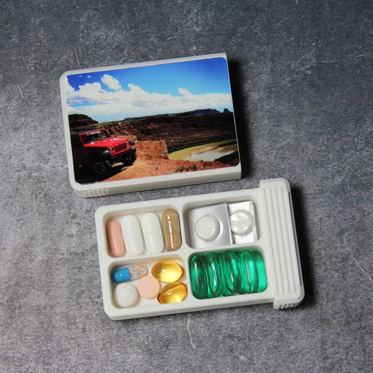 Custom photo pill organizer, white, lid removed: 4-compartment base with double cells holding tablets, a cut blister pack and green softgels