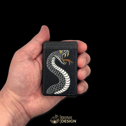 Pilulier édition artistique Snake Tattoo fermé, tenu en main pour donner l'échelle, serpent argenté, crocs et œil dorés, langue fourchue cuivre.