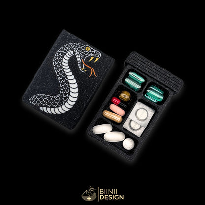 Pilulier édition artistique Snake Tattoo : serpent argenté aux crocs et œil dorés sur galaxy black, à côté d'une base 5 compartiments remplie.