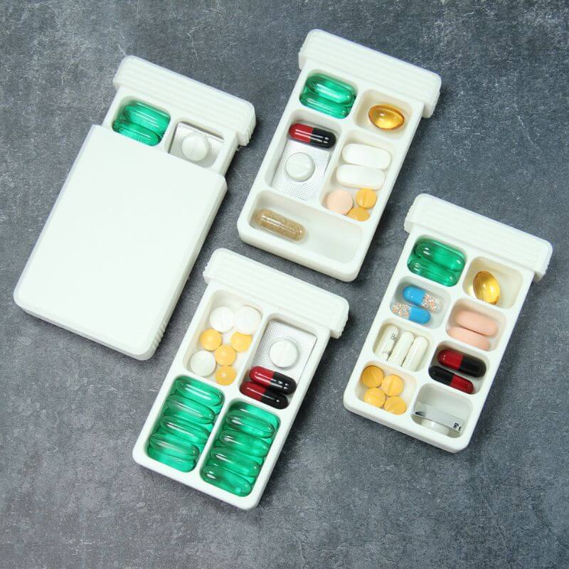 Four white travel pill organizers on slate: one closed, three with the lid slid open showing four to eight compartments filled with capsules, tablets and a cut blister.