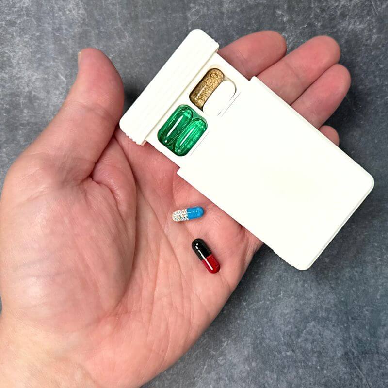 White travel pill organizer with the lid slid open in an open palm, two compartments filled with capsules, two more capsules resting on the hand.