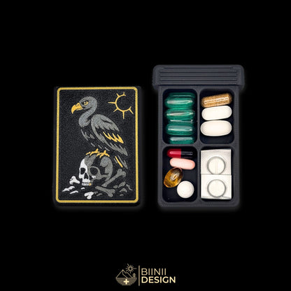 Vulture's Shadow art edition pill organizer lid beside its 4-compartment base filled with assorted supplements, vulture and skull artwork.