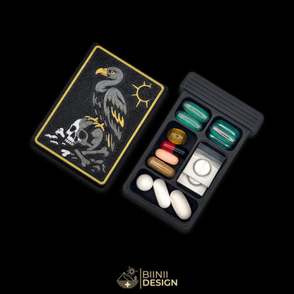 Vulture's Shadow art edition pill organizer: anthracite vulture above a silver skull half in its shadow, on matte black, beside a filled 5-compartment base.