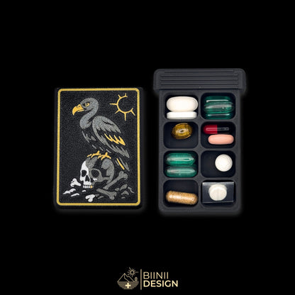 Vulture's Shadow art edition pill organizer lid beside a filled 8-compartment base holding tablets, capsules and supplements.