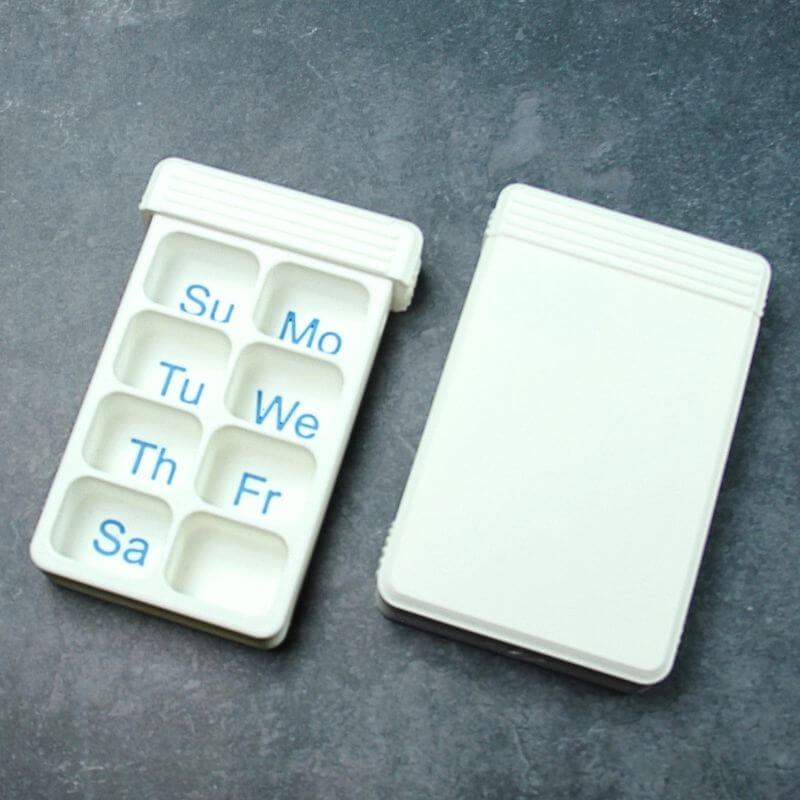 White compact 7-day pill organizer open with blue day letters Su to Sa in seven compartments, next to a second one closed, on slate.