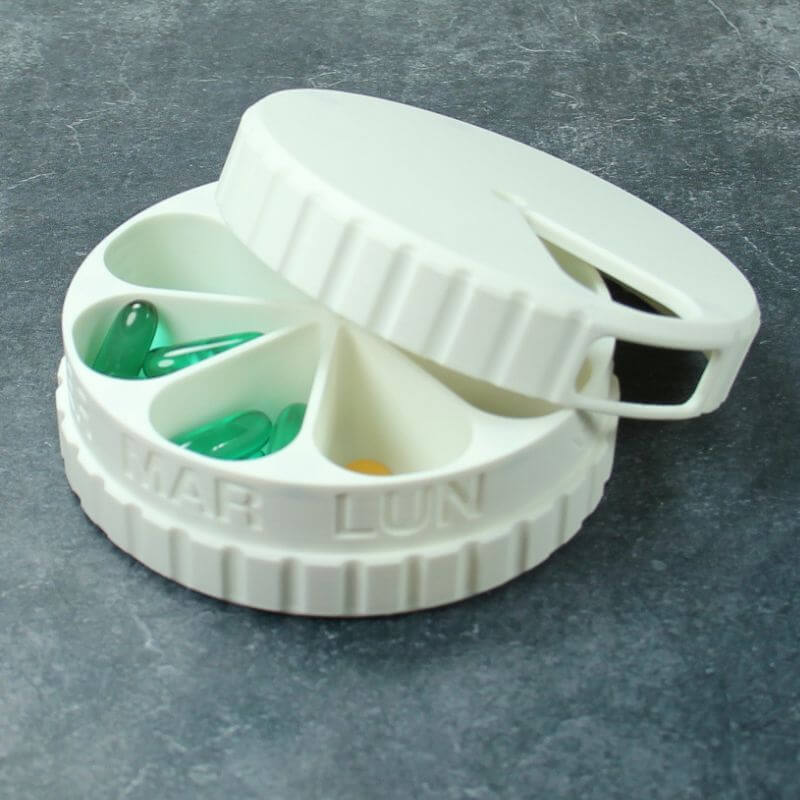 White round weekly pill organizer with the lid turned open over the Monday and Tuesday compartments, embossed LUN and MAR, holding green capsules, on slate.